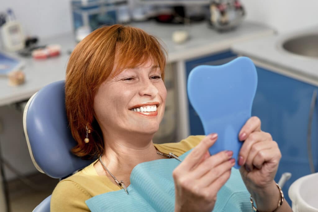 Smiling senior woman with new dental implants sitting in the dental office and looking at the mirror
